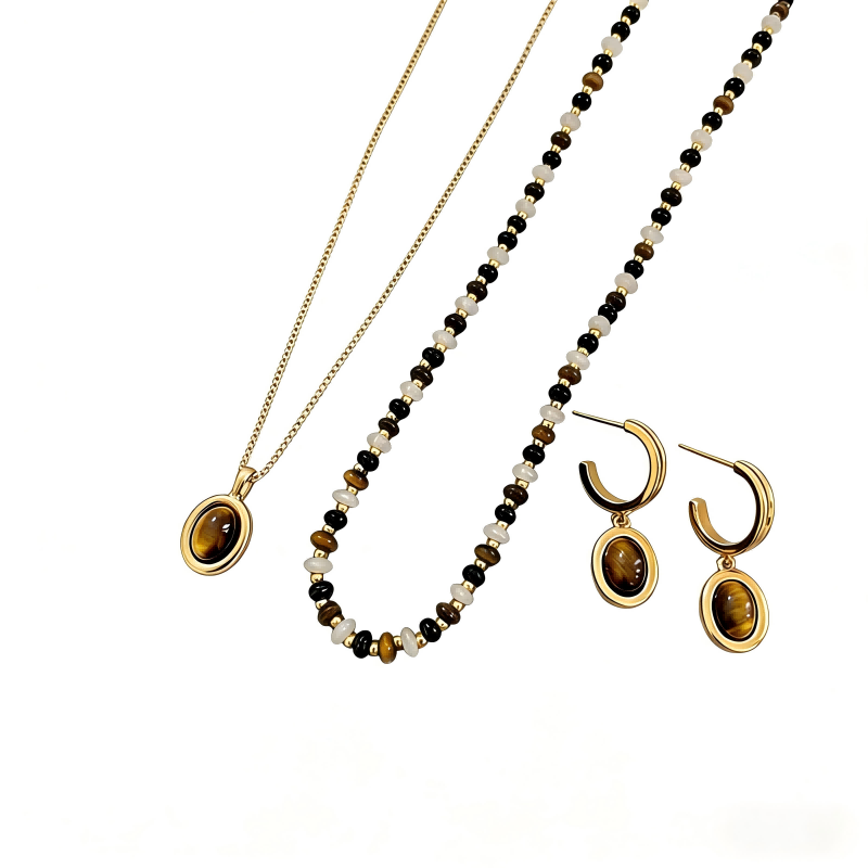 A gold tiger's eye jewelry set displayed on a white background. The collection features a delicate gold chain necklace with an oval tiger's eye pendant, a beaded necklace with alternating brown, black, and cream stones, and a pair of gold hoop earrings with matching tiger's eye drop charms.