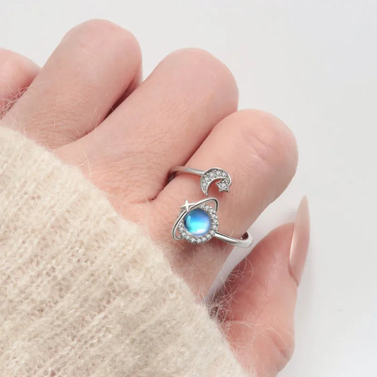 Mercury brand stainless steel spinning ring on a woman’s hand. The lifestyle photo features a rotating blue moonstone planet and crystal crescent moon design, paired with a soft cream knit sweater for a cozy, elegant aesthetic.